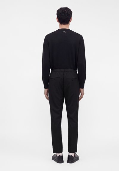 Homme debout, dos à la caméra, portant une chemise noire à manches longues, un pantalon noir avec des poches arrière, des chaussettes blanches et des chaussures noires sur fond blanc.