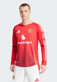Maillot de football à manches longues rouge avec des rayures blanches, arborant le logo de Manchester United et le logo "Snapdragon", fabriqué en matériau évacuant l'humidité.