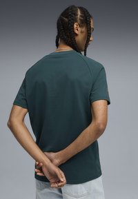 Junger Mann mit geflochtenem Haar steht mit dem Rücken zur Kamera, trägt ein dunkelblaues T-Shirt und helle Jeans, die Hände hinter dem Rücken gefaltet.