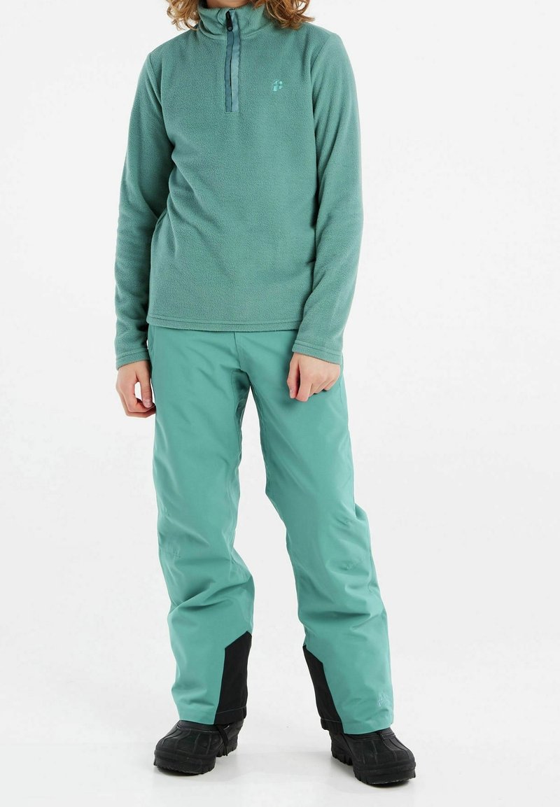 Pull polaire vert clair avec col demi-fermeture éclair, associé à un pantalon imperméable bleu sarcelle avec des accents noirs et des bottes noires robustes.