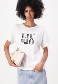 Balta medvilninė marškinėliai su trumpomis rankovėmis ir apvaliu kaklu, ant kurių juodai užrašyta "LIU JO". Modelis laiko šviesiai smėlinę rankinę.