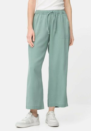 Persoon die lichtgroene wijde broek met trekkoord en witte sneakers draagt, staand tegen een effen witte achtergrond.