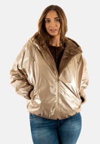 Chaqueta bomber metálica beige con cuello de piel sintética, cierre frontal con cremallera y puños fruncidos, llevada sobre un suéter oscuro y jeans azules.