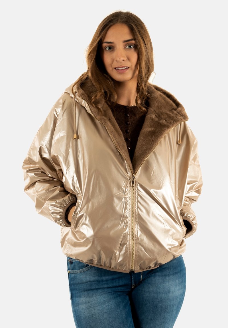 Chaqueta bomber metálica beige con cuello de piel sintética, cierre frontal con cremallera y puños fruncidos, llevada sobre un suéter oscuro y jeans azules.
