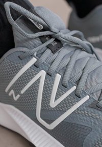 Grå atletiske sko med en tekstureret mesh-overdel, hvid branding og snørebånd. Sålen er hvid med udtalt mønster for trækkraft.