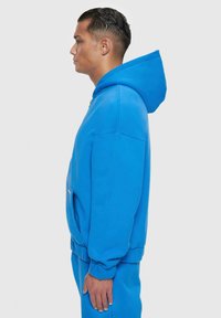 Blauer Hoodie mit großer Kapuze, seitlichen Taschen und gerippten Bündchen. Aus weichem Stoff mit glatter Textur, im Seitenprofil gezeigt.