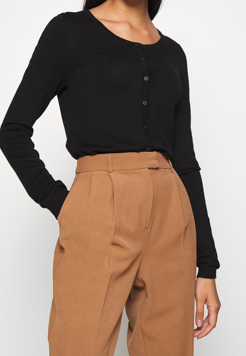 Femme portant un haut noir cintré à manches longues boutonné, rentré dans un pantalon camel brun plissé taille haute, les mains dans les poches.