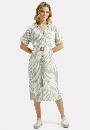 Kobieta stoi z rękami za plecami, ubrana w plisowaną, zapinaną na guziki, midi sukienkę z krótkim rękawem w zielono-biały wzór liści oraz białe trampki.