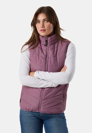 Jeune femme aux cheveux bruns portant un gilet matelassé mauve sur une chemise blanche à manches longues et un jean bleu, bras croisés, fond neutre.
