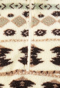 Close-up van een crèmekleurige en bruine gestreepte fleece met getextureerde, geometrische tribale patronen en een centrale rits.