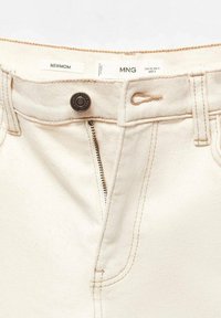 Gros plan sur un jean couleur crème avec braguette partiellement dézippée, bouton en métal et étiquettes visibles portant les inscriptions "NEWMOM" et "MNG".