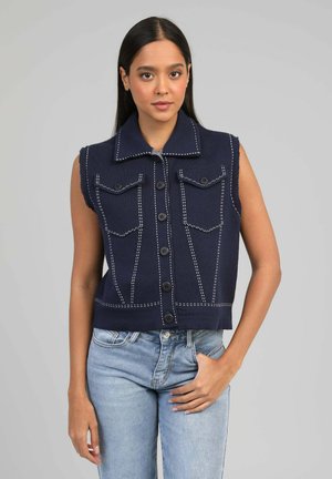 Jeune femme aux longs cheveux noirs portant un gilet sans manches bleu marine à boutons avec des coutures blanches et un jean bleu clair.