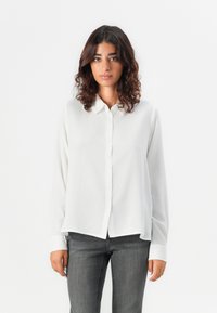Camicia bianca con bottoni, dal tessuto liscio, maniche lunghe e orlo arrotondato, abbinata a jeans aderenti grigio scuro. Design semplice e minimalista.