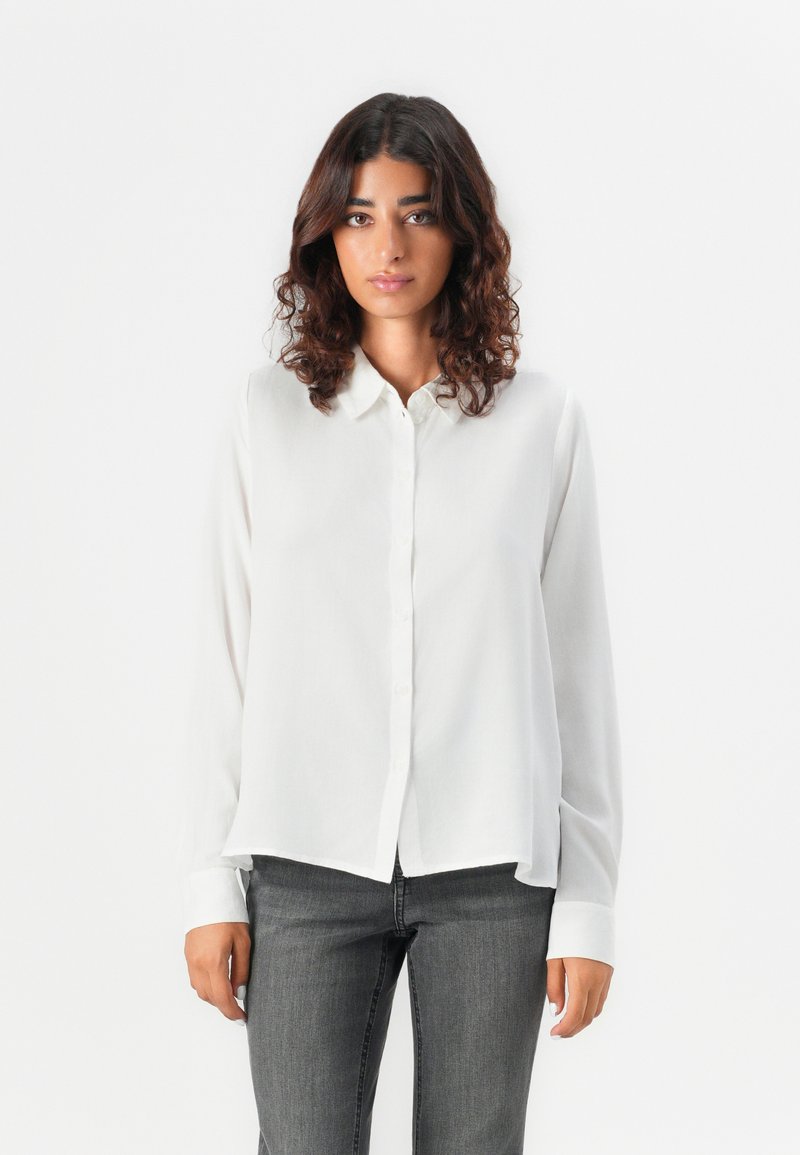 Camicia bianca con bottoni, dal tessuto liscio, maniche lunghe e orlo arrotondato, abbinata a jeans aderenti grigio scuro. Design semplice e minimalista.