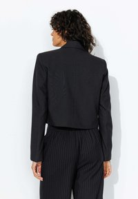Blazer noir cintré avec des manches longues, une texture subtile et un col cranté ; porté sur un pantalon noir à rayures.