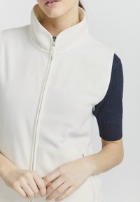 Weißer Fleece-Weste mit hohem Kragen und Frontreißverschluss. Enthält strukturierte marineblaue Ärmel. Glattes Material mit seitlichen Taschen und sauberen Nähten.
