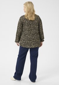 Blusa a maniche lunghe con motivo nero e verde oliva, abbinata a jeans larghi blu scuro e calzature di colore chiaro.