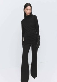 Jersey de cuello alto negro combinado con pantalones acampanados negros de talle alto. La modelo lleva guantes largos negros, presentando un look elegante y minimalista.