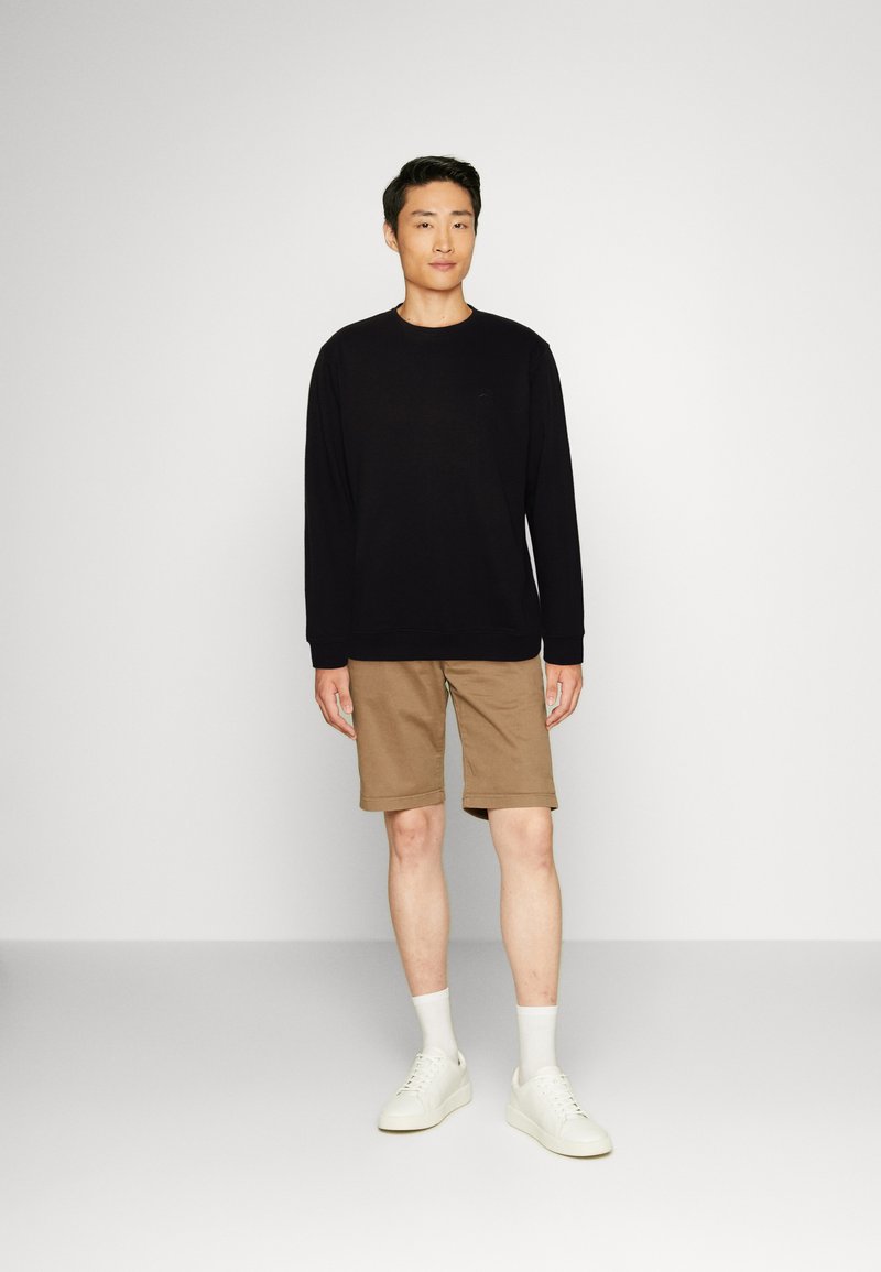Maglietta nera a maniche lunghe, pantaloni corti beige, calzini bianchi e sneaker bianche. Il modello è in piedi contro uno sfondo neutro.