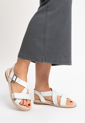 Feet wearing white flat sandals with multiple straps and a buckle, paired with a dark ankle-length skirt, toes painted red.