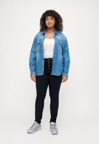Camisa de denim en azul claro, abotonada, con dos bolsillos en el pecho. Llevada sobre una camiseta blanca, combinada con pantalones negros de cintura alta y zapatillas plateadas.