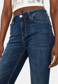 Jeans in denim blu scuro a vita alta, con una texture liscia, chiusura con bottone e cuciture visibili lungo i lati e le tasche.