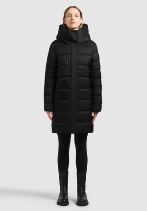 Mujer con un abrigo de invierno largo y acolchado de color negro con cuello alto, leggings negros y botas negras hasta el tobillo, de pie frente a un fondo liso.
