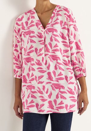 Kobieta ubrana w białą tunikę-bluzkę z różowym kwiatowym wzorem, dekolt w serek, rękawy 3/4 z elastycznymi mankietami, w połączeniu z ciemnoniebieskimi dżinsami.