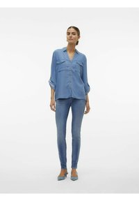 Camicia di jeans azzurro chiaro con chiusura a bottoni, due tasche sul petto e maniche arrotolate, abbinata a jeans attillati azzurro chiaro e tacchi.
