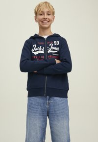 Felpa con zip blu navy con testo del logo; materiale in cotone, maniche lunghe e tasche frontali. Abbinata a pantaloni shorts in denim azzurro.