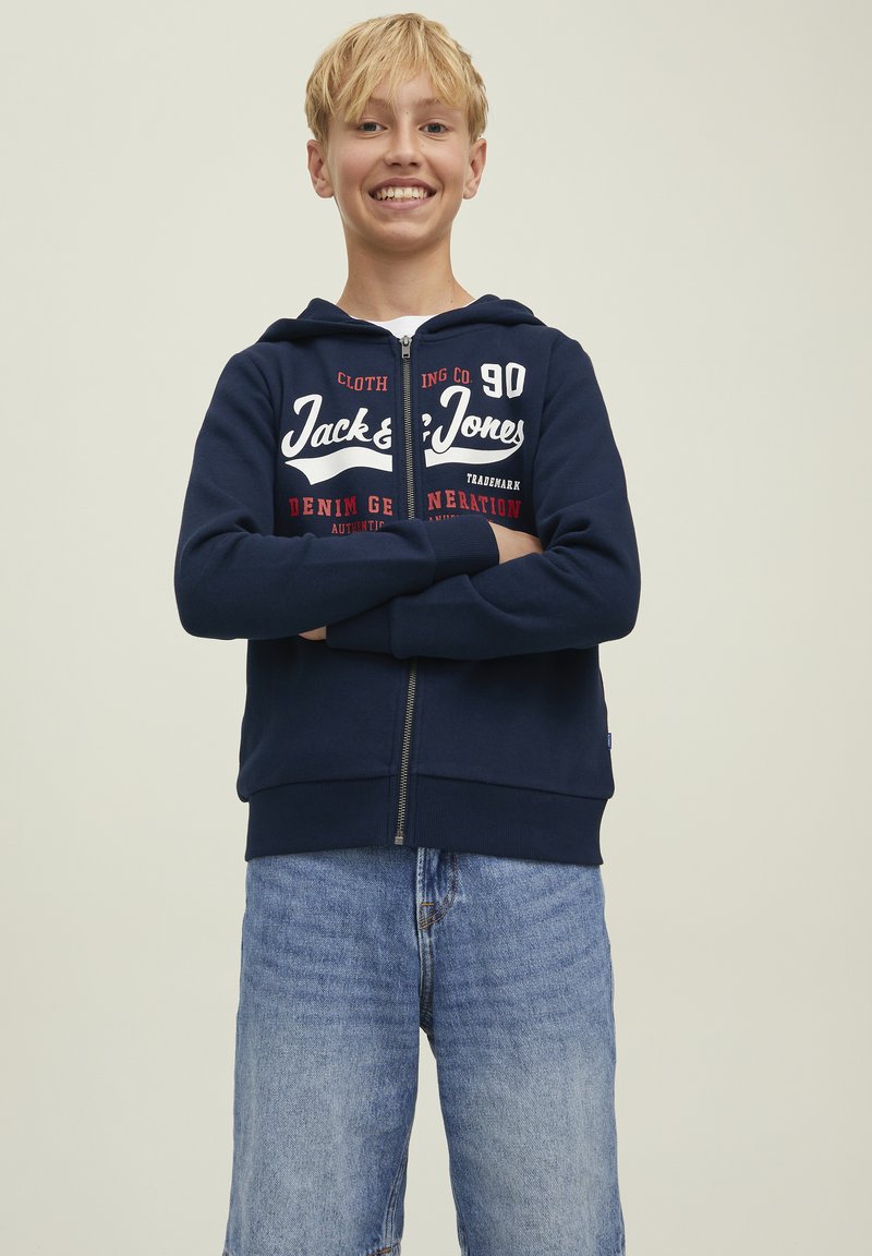 Felpa con zip blu navy con testo del logo; materiale in cotone, maniche lunghe e tasche frontali. Abbinata a pantaloni shorts in denim azzurro.