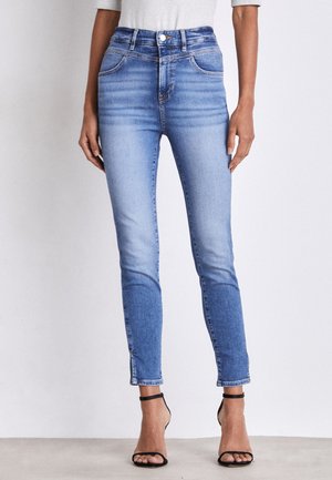 Femme portant un jean skinny taille haute bleu clair avec des détails délavés et des sandales à talons hauts noires avec des brides, debout devant un fond uni.