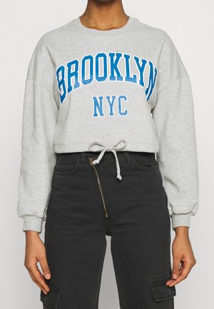 Person iført en lysegrå cropped sweatshirt med blå "BROOKLYN NYC" tekst og højttaljede sorte bukser med en vinklet lynlås.
