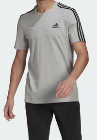 Camiseta gris de manga corta con diseño de tres rayas negras en los hombros y pequeño logo negro en el pecho, hecha de una mezcla de algodón.