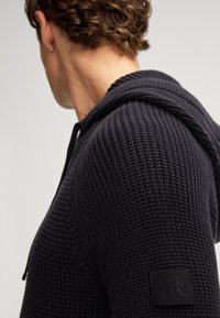 Czarna, strukturalna bluza z kapturem i ściągaczami, z logo naszytym po lewej stronie. Tkanina ma wzór dzianiny.