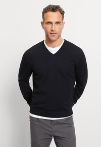 Zwart gebreide trui met v-hals, gedragen over een wit shirt met ronde hals, met lange mouwen en een aansluitend ontwerp. Grijze broek is zichtbaar.