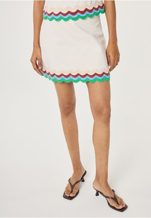 Woman wearing a cream skirt with scalloped edges in tan, red, blue, and green, paired with black kitten-heeled sandals.