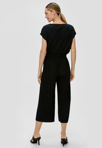 Zwarte geribbelde jumpsuit met korte mouwen, elastische taille en brede cropped benen. G gedragen met zwarte hakkenandalen, weergegeven van achteren.