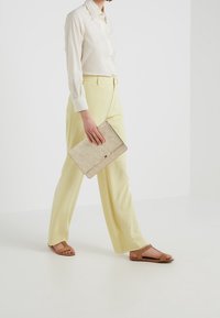 Beige Clutch mit strukturierter Oberfläche, in einer Hand gehalten. Das Outfit besteht aus einem weiß gestreiften Hemd und hellgelben Hosen, kombiniert mit braunen Sandalen.