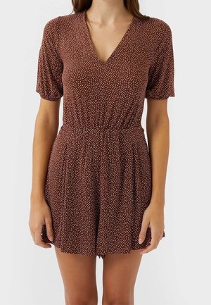 Femme portant une combinaison courte à manches courtes marron à col en V avec des pois blancs et une ceinture élastique, sur un fond blanc uni.