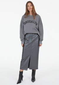 Gray sweatshirt with "BROOKLYN" text, paired with a high-waisted, glossy, dark gray leather skirt featuring a side slit and pockets. Black heeled boots.