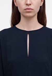 Gros plan d'une femme portant un haut bleu marine avec une petite fente verticale sous l'encolure ronde, affichant une expression faciale neutre.