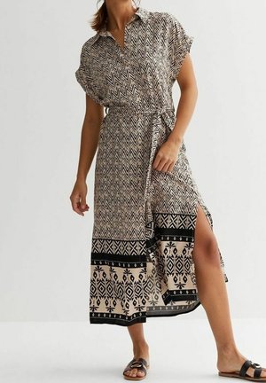 Robe longue à motif chevron en beige et noir, manches courtes, col, ceinture à nouer et fente latérale. Portée avec des sandales noires.
