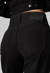 Vaqueros de denim negro con una textura suave, con un parche de cuero en la cintura y dos bolsillos traseros con costuras decorativas.