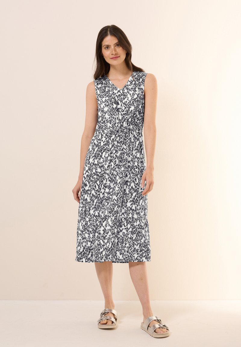 Ärmelloses V-Ausschnitt-Kleid mit einem schwarz-weißen Muster, tailliertem Schnitt, knielang und aus leichtem Stoff. Kombiniert mit metallischen Sandalen.