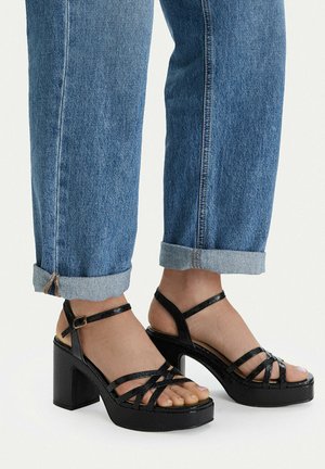 Pies calzando sandalias negras de tacón alto con tiras y tacones gruesos, combinadas con jeans azules arremangados sobre un fondo claro y liso.