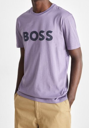 Homme portant un t-shirt violet clair avec un grand texte noir "BOSS", associé à un pantalon beige, mains dans les poches, sur un fond uni.