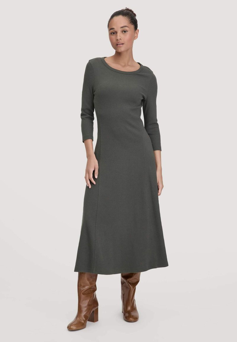 Frau in einem olivgrünen, langärmligen Midikleid mit braunen, hohen Lederstiefeln, stehend vor einem schlichten, hellen Hintergrund.