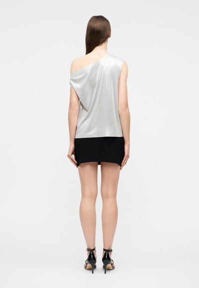 Γυναίκα που στέκεται με την πλάτη στραμμένη, φοράει ένα ασημί τοπ με έναν ώμο, μαύρη φούστα και μαύρα σανδάλια με ψηλό τακούνι σε απλό φόντο.