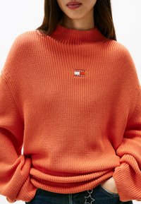 Maglione arancione lavorato a maglia con un colletto alto a coste. Presenta un piccolo patch logo "Tommy Jeans" rosso e bianco sul davanti.